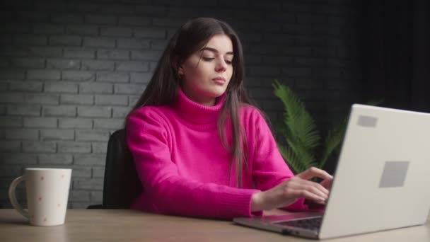 一个笑着的小女孩坐在工作室里 正在笔记本电脑上打字 一个穿着粉色毛衣的女孩在笔记本电脑上远程工作 高质量的4K镜头 — 图库视频影像