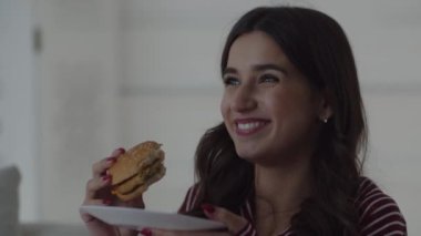 Güzel bir gülüşü olan, hamburger yiyen genç bir kadının videosu. Genç bir kız televizyonun önünde yemek yiyor. Bir kadın televizyon izlerken güler. Yüksek kalite 4k görüntü