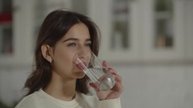 Beautiful woman in a white sweater drinking water from a glass. A brunette girl smiles after quenching her thirst. Pretty young lady takes a sip. Hydration concept. High quality 4k footage