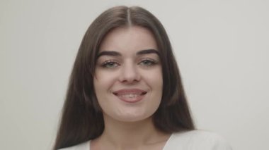 Young girl on a white background smiling while looking at the camera. Front view of attractive brunette woman blinking with smile. High quality 4k footage
