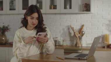 A brunette woman sitting in her home office uses her phone to search for information that will help her solve a work task. Remote work using phone and laptop. Girl scrolling on smartphone screen. High