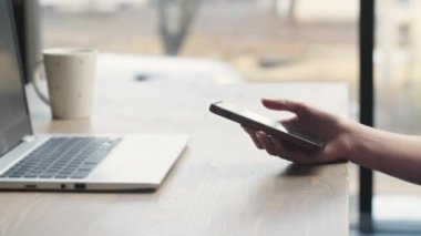 Girl freelancer sitting in a cafe uses the phone for remote job. Work from cafe using laptop and phone. Female hand scrolling smartphone screen. High quality 4k footage