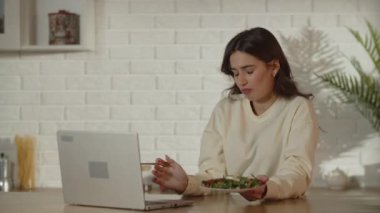 Kadın bir serbest çalışan iş sırasında salata yiyor. Duygusal kadın dizüstü bilgisayarında videolar izliyor ve yemek yiyor. Esmer kız mutfakta öğle yemeğinin tadını çıkarıyor ve bilgisayar ekranına bakıyor. Yüksek kalite