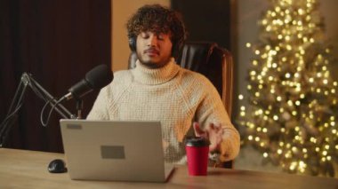 Online influencer is doing podcast. Smiling young man in headphones streaming on Christmas and drinking coffee. High quality 4k footage