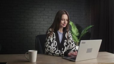 Smiling female freelancer is typing fast on her laptop. A brunette woman works with computer at home office. Cheerful girl in cardigan enjoying distant job. High quality 4k footage