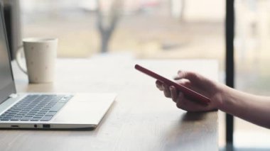 A freelancer girl uses a phone and a laptop to solve work tasks. Cropped view of woman working from cafe. Female hand typing and scrolling on smartphone. High quality 4k footage