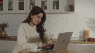 Video of a gloomy woman closes her laptop and sips coffee from a white cup. A tired brunette girl taking a coffee break. Exhausted freelancer finishing work in kitchen. High quality 4k footage