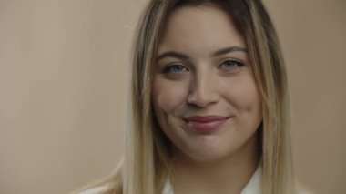 Close up portrait of smiling blonde woman. Front view of blue-eyed girl looking at camera with smile and blinking on beige background. High quality 4k footage