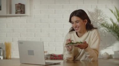 Esmer bir kadın, dizüstü bilgisayarında video izlerken salata yiyip gülüyor. Freelancer dersleri sırasında ara verip öğle yemeği yemeye karar verdi. Mutfakta yemek yerken gülümseyen kız.
