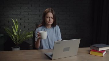 A sleepy woman drinks coffee from a cup to cheer herself up and finish a work project. Video of a woman yawning while working on a laptop. High quality 4k footage