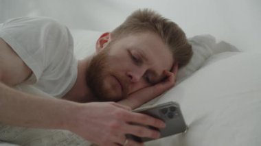 Man setting alarm on his phone before taking a nap. European guy checking his messages and falling asleep in bed. High quality 4k footage