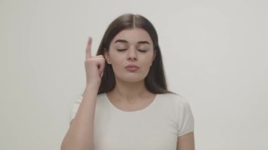 A young brunette with a serious look taps her finger on her temple and points in the direction of the camera. Studio shot of a girl in a white T-shirt on a white background. High quality 4k footage