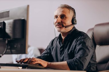 Sales manager talking to client via headset. Middle-aged hotline operator typing on computer keyboard. Customer support agent in office. High quality photo