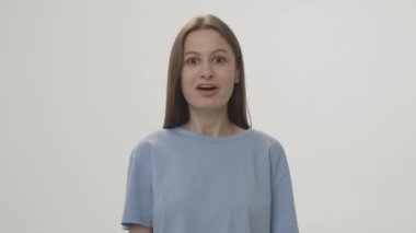 The brunette looks directly into the camera and shows thumbs up. Studio shot of a woman against a white wall. High quality 4k footage