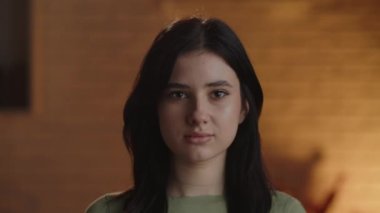 The brunette woman raises her eyes and looks to the camera with a slight smile on her lips. Portrait shot of a smiling girl against a blurred background. Front view of shy young lady. High quality 4k