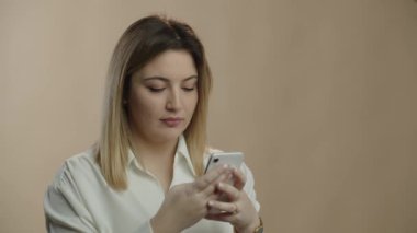 Pretty young woman typing message and looking away with smile. Studio footage of blonde girl with smartphone smiling on brown background. High quality 4k footage