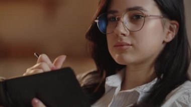 Gözlüklü genç bir iş kadını defterine görevlerin bir listesini yazar. Esmer kız notlar alıyor. Odaklanmış yüz ifadesiyle çalışan kız öğrenci. Yüksek kalite 4k görüntü