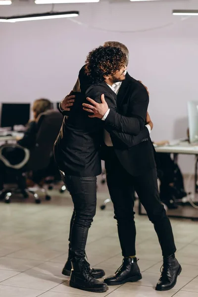 Smiling businessmen hugging in the office. Coworkers in suits embrace each other. Full length view of managers celebrating successful deal. High quality photo