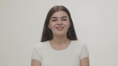 Güzel bir esmer gözlerini açar ve doğrudan kameraya bakarken gülümsemeye başlar. Beyaz tişörtlü neşeli bir esmerin stüdyo fotoğrafı. Yüksek kalite 4k görüntü