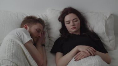 A married couple sleeps on the bed, but the wife wakes up from the sound of the alarm clock and wakes up the husband. Video in which a married couple wakes up due to the alarm clock going off. High