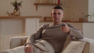 A young man sitting on an armchair and wrapped in a blanket switches channels on the TV. A man spends the evening watching TV. High quality 4k footage