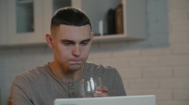 A man with a short haircut is working on a laptop and drinking water from a glass. Freelancer works remotely at a computer and takes a sip from a water glass. High quality 4k footage