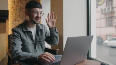 The young brunette man joyfully greets the interlocutor during online conversation. Freelancer in glasses talks via video call. Stylish guy with laptop having team conference in cafe. High quality 4k