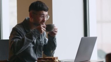 Freelancer drinking coffee while working in cafe. Smiling young man in glasses using laptop in coffeehouse. Cheerful guy typing on computer keyboard in the restaurant. High quality 4k footage