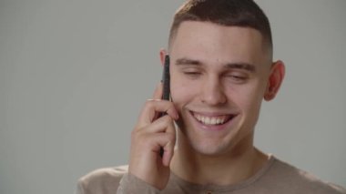 Telefonda konuşurken kısa saçlı, gülümseyen genç bir adamın yakın çekimi. Genç bir adam telefonda bir şey üzerinde çalışıyor. Beyaz bir arka planda stüdyo çekimi. Yüksek kalite 4k
