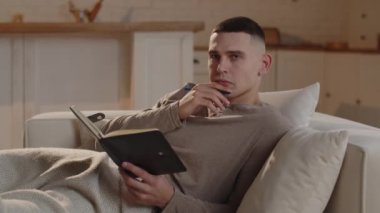 Kanepede uzanmış kısa saçlı bir adam gelecek hafta sonu için yapılacakların bir listesini yapar ve kâğıt deftere yazar. Bir adam düşüncelerini siyah bir deftere yazar.