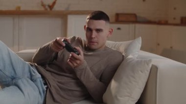 Bir video oyununda zor bir seviyeyi geçmeye çalışan bir adamın elinde oyun platformuyla kanepede yattığı yakın çekim. Genç bir adam video oyununun son patronunu atlattıktan sonra sevinç duyar. Yüksek