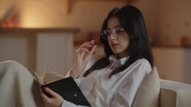 一位女自由职业者躺在沙发上 列了一张待办事项清单 一个戴眼镜的年轻的黑发女人在笔记本上写笔记 高质量的4K镜头 — 图库视频影像