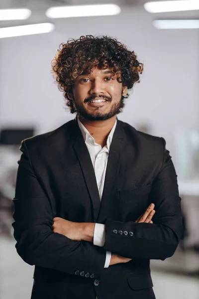 Cheerful young businessman posing with folded arms. Successful entrepreneur with curly hair looking at camera with smile. Happy Indian manager in black suit. High quality photo