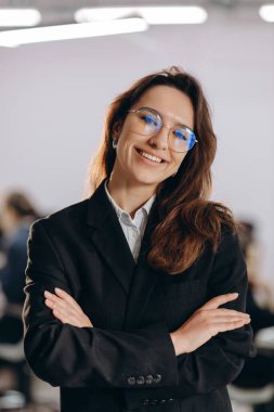 Attractive businesswoman in glasses posing with folded arms. Indoor shot of smiling young woman in black suit standing in office. High quality photo
