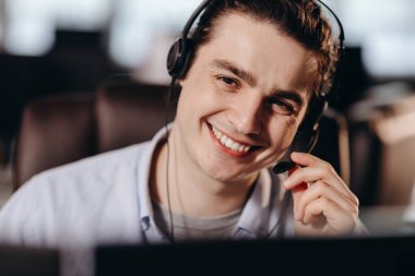 Smiling male receptionist in headphones. Happy call center operator in headset on the blurred background. Indoor shot of online consultant. High quality photo