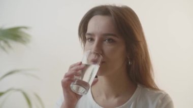 Genç bir kadın bir bardak sudan bir yudum alıyor. Kahverengi gözlü sevimli bir kız su içtikten sonra kameraya bakar. Kadın güne içme suyuyla başlıyor. Yüksek kalite 4k görüntü