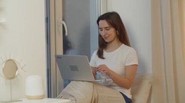 Gülümseyen kadın, arkadaşlarıyla dizüstü bilgisayar kullanarak sohbet ediyor. Bir kız odasındaki pencere eşiğinde oturuyor ve bilgisayar kullanıyor. Evden çalışıyorum. Yüksek kalite 4k görüntü