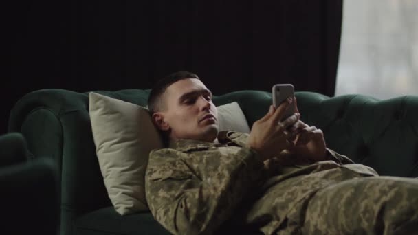 一个身穿军服的年轻人躺在沙发上 翻动着他的智能手机 一个年轻的士兵正在用手机浏览新闻 等待着一位心理医生的到来 — 图库视频影像