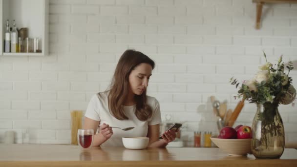 朝食をとりながらソーシャルメディアのフィードをスクロールする女性のビュー 白いTシャツを着た女の子がキッチンのテーブルに座り 白いボウルからお粥を食べています 高品質4K映像 — ストック動画