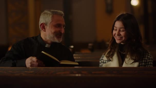 祭司は聖書を手にして教区の人と話をする教会に座っています 日曜日の礼拝の後 カトリックの司祭は笑顔のブルネットの女性と通信します 高品質4K映像 — ストック動画