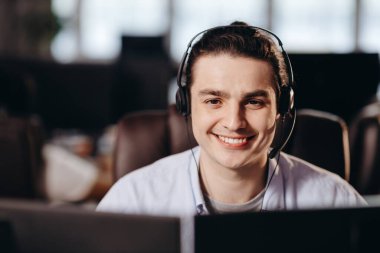 Kapalı alanda, kulaklıklı gülümseyen yardım hattı operatörünün fotoğrafı. Kulaklıklı genç müşteri destek ajanı bilgisayar ekranına bakıyor. Yüksek kalite fotoğraf