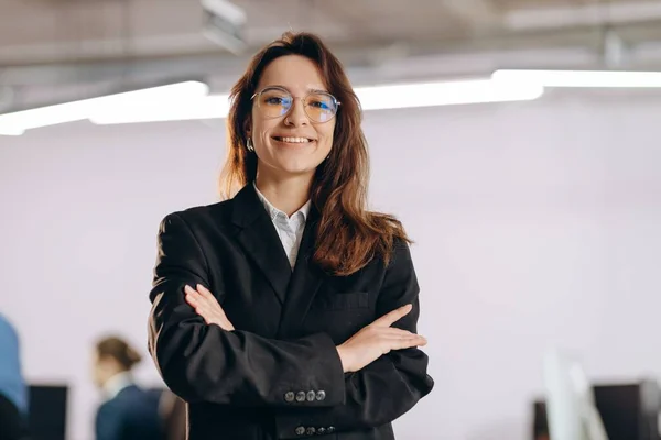 Female top manager posing in office with smile. Happy businesswoman standing with folded hands in office. Successful woman in formal wear. High quality photo