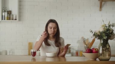 Kahvaltıda yulaf ezmesi yiyen bir kadının videosu. Mutfakta oturup telefonundan bir şeye bakarken. Esmer bir kadın kahvaltı yaparken telefonundan komik videolar izliyor. Yüksek kalite 4k