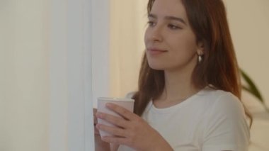 Kız pencereden manzaraya hayran ve beyaz bir fincandan çay içiyor. Çay içip kameraya gülümseyen genç bir kadının videosu. Yüksek kalite 4k görüntü