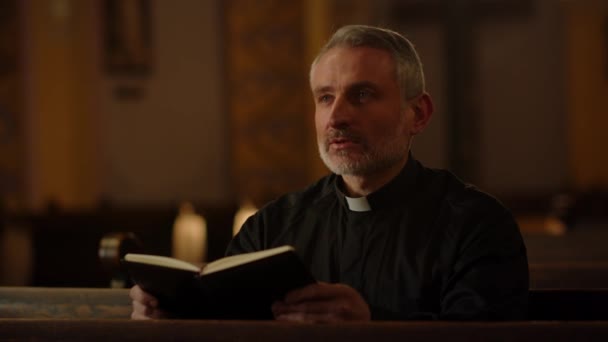一位灰白头发的天主教牧师坐在教堂的长椅上 手里拿着一本圣经 一位读圣经的天主教牧师的近照 高质量的4K镜头 — 图库视频影像