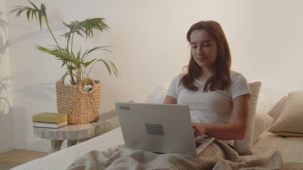若いブルネットの女性はベッドに横たわっている間ラップトップを使用します 白いTシャツを着た女の子が膝の上にノートパソコンのキーボードを押している ベッドで働いてるフリーランス 内務省だ 高品質4K映像 — ストック動画
