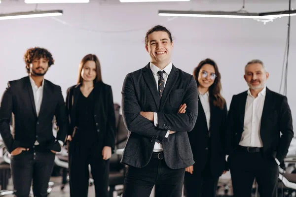 Young business leader posing with smile in front of colleagues. Happy businessman and his coworkers standing in office. High quality photo