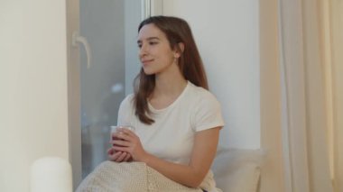 Pencerenin kenarında bir fincan sıcak çayla oturan esmer bir kadın görüntüsü. Beyaz tişörtlü bir kadın pencerenin manzarasına hayran. Pencere kenarında oturmuş elinde bir fincan kahve tutuyor.