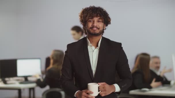ビジネススーツを着た若いインド人男性がコーヒーを飲んでカメラを見ている ぼやけたオフィスの前で笑顔の男性ビジネスマンのビュー 高品質4K映像 — ストック動画
