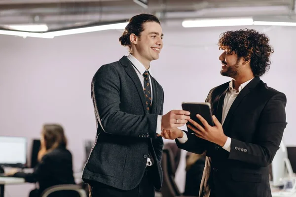 タブレットを見てお互いに笑顔のビジネスマン 正式な着用で幸せなオフィスの労働者の屋内ショット デジタルタブレットを使用して2人の起業家 高品質の写真 — ストック写真